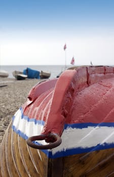 An upturned wooden constructed small fishing boat, resting on a pebbled beach, in the midst of being repaired and painted for the fothcoming fishing season. The bottom of the boat being painted in red with a blue stripe. The union jack flag flies in the background.