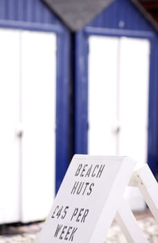 A hand painted sign advertising beach huts for hire. Blue and white beach huts visible in soft focus to background. Located at Beer, in Dorset, England.