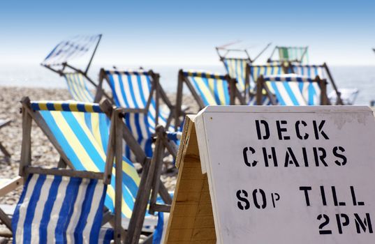 A landscape format image of a hand made advertising board, advertising beach side deck chairs for hire. Various colored deck chairs in soft focus to background. Located in Beer in Dorset, England.