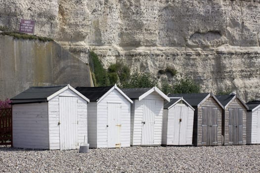 A landscape format image of a row of wooden beach huts set against chalk cliffs. Located on the jurassic coast line in Beer, Dorset, England.