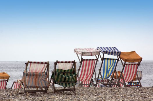 A row of various colored striped deck chairs with canopys, on a pebbled beach. Located in Beer in rural Dorset, England. Viewed from behind with the sea and blue sky visible to the background.