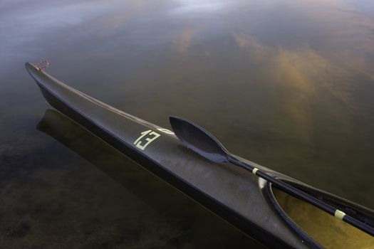 bow of sea racing kayak (lightweight and slim carbon kevlar fiber design) with a wing paddle across cockpit on calm water with cloud reflections, temporary racing number 13 on front deck, wet after paddling

l

eightweight carbon kevlar racing sea kayak with wing paddle on a front deck 