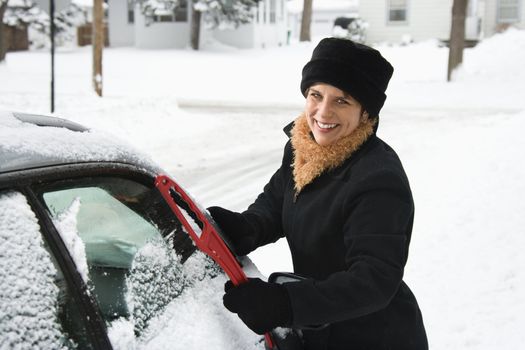 Caucasian mid adult woman scraping ice off car windshield and smiling at viewer.