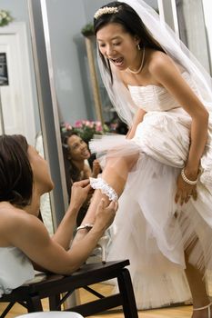 African-American bridesmaid placing garter on Asian bride's leg.