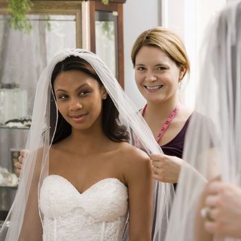 Caucasian seamstress helping African-American bride with veil in bridal shop.