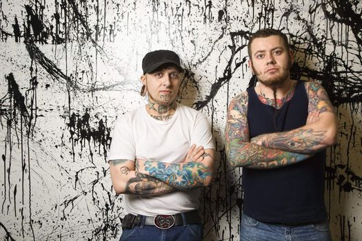 Two Caucasian tattooed men standing against paint splattered background.