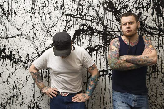 Two Caucasian tattooed men standing against paint splattered background.