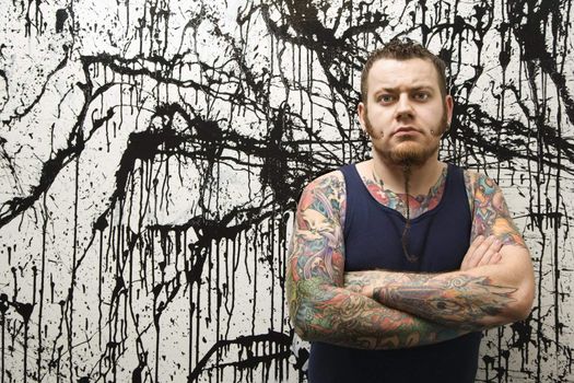 Caucasian tattooed man with braided beard standing against paint splattered background.