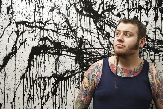 Caucasian tattooed man with braided beard standing against paint splattered background.