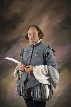 William Shakespeare in period clothing holding feather pen standing and looking at viewer.