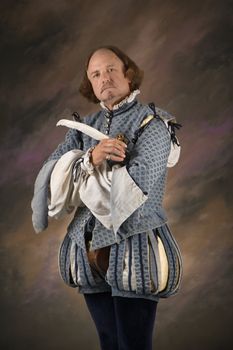 William Shakespeare in period clothing holding feather pen and looking at viewer.