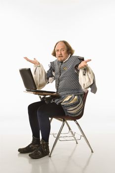 William Shakespeare in period clothing sitting in school desk with laptop and shrugging at viewer.