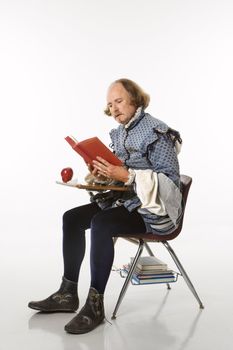 William Shakespeare in period clothing sitting in school desk reading a book.