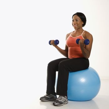 African American young adult woman sitting on exercise ball holding dumbbells.