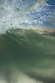 Underwater view of a curling, breaking wave; With the bright blue sky and sunset on the horizon coming through the surface and the stirring sand on the ocean floor