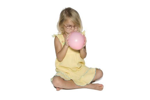 A young girl in a yellow dress sitting down and blowing up a pink balloon.  Isolated on a white background