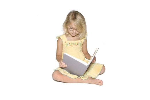 A young blonde girl sitting crosslegged while reading a textbook.  Isolated on a white background