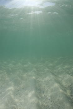 Underwater view of sunbeams stream through the surface of the ocean and dancing on the floor of the sea on a bright sunny day.  Bright sand and blue sky.