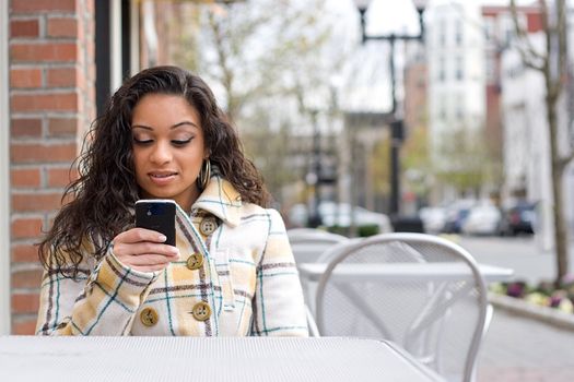 An attractive Indian woman texting or searching the web on her cell phone while seated at a table outdoors.