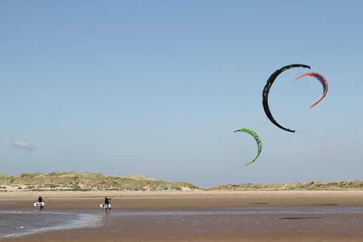 People walking kites along the beach carrying boards.