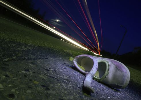 Night shot of a child's shoe after a car accident with headlight streams zipping through the frame.