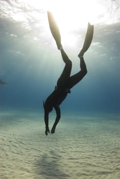 A freediver descends to the ocean floor as the sun streaks beams of light from the surface of the ocean