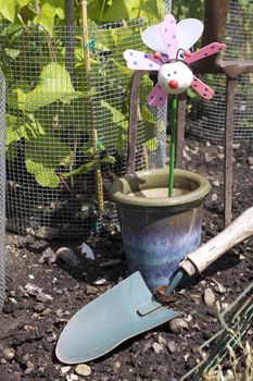 A childs plant pot decoration of a stylised bee. Set in a blue glazed ceramic plant pot in a vegetable garden amongst growing runner beans. A small hand held garden trowel lies in front of the pot.