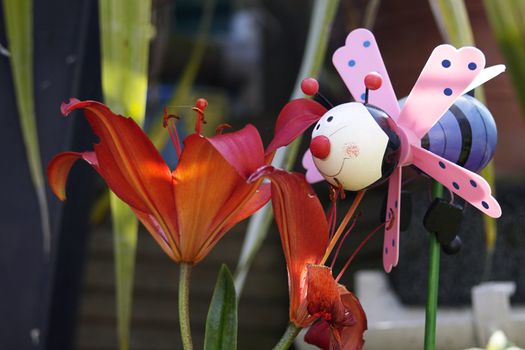 A childs plastic plant pot toy decoration of a stylised bee with a bright blue body and pink wings. Set in a garden pot pointing towards and sniffing at bright orange flowers.