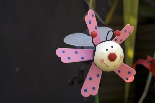 A childs plastic plant pot toy decoration of a stylised bee with pink wings. Set in a garden flower pot.