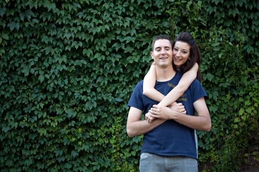 A happy young couple in their mid 20s together outdoors in front of some green ivy.