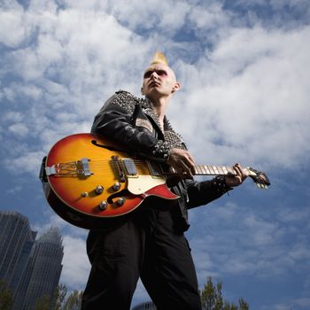 Portrait of mid-adult Caucasian male punk holding guitar with skyline in background.