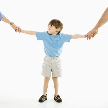 Boy holding hands with parents standing against white background.