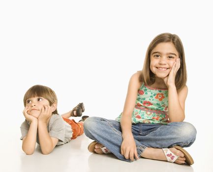 Girl sitting smiling next to boy lying on stomach looking bored.