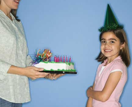 Girl wearing party hat with mother holding birthday cake.