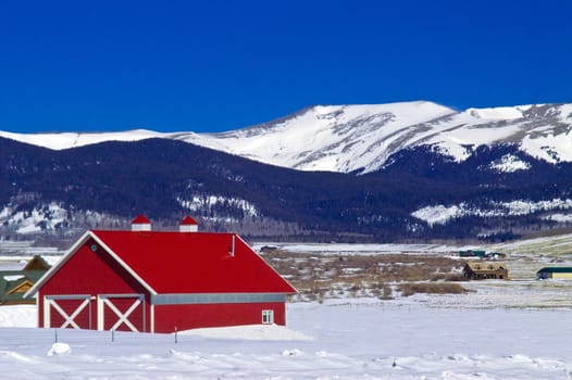 A classic red barn in Colorado winter with snow capped mountain peaks, portrays rural America