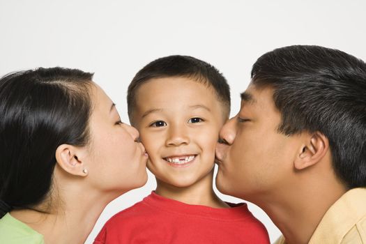 Asian mother and father kissing opposite cheeks of smiling son in front of white background.