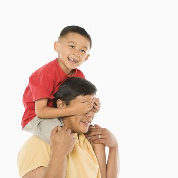 Asian boy sitting on father's shoulders with hands over his eyes in front of white background.