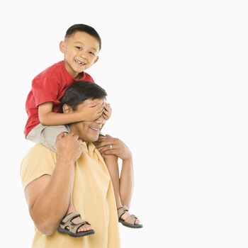 Asian boy sitting on father's shoulders with hands over his eyes in front of white background.