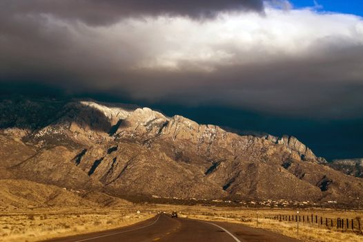 A highway in New Mexico showing the Sandia Mountains seems to lead to the awaiting thunderstorms in evening light just before sunset