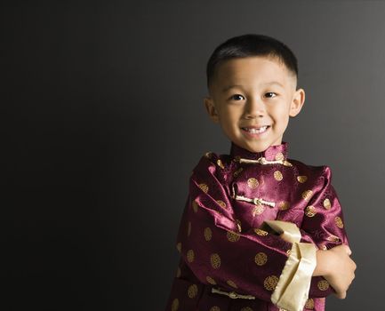 Asian boy in traditional attire standing against black background.