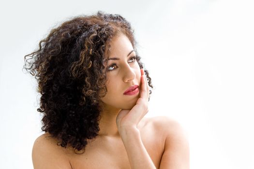 Sincere beautiful young woman with brown curly wild hair and bare shoulders with hand on chin, isolated