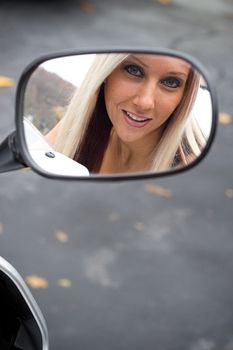 A pretty blonde woman looking into the mirror on her motorcycle.