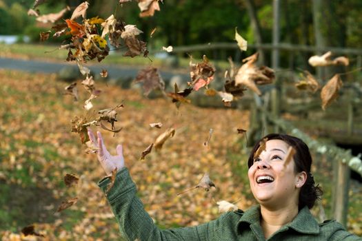 A pretty young woman laughing as the leaves fall all around her during the autumn season.