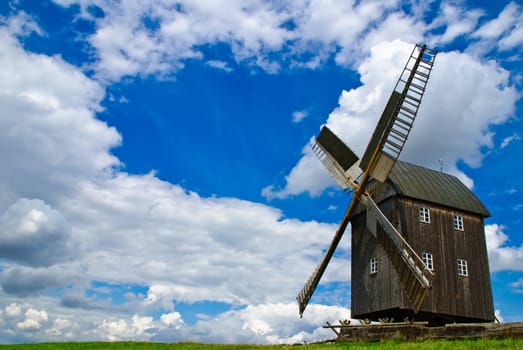 Wooden windmill against the summer blue sky with white clouds on a green hill with a birch