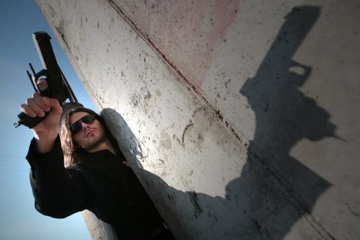 man in black sunglasses and with gun peers because of the concrete wall