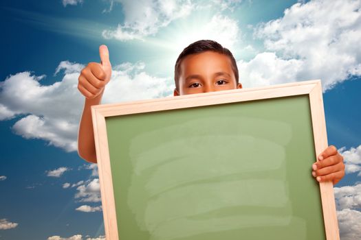 Proud Hispanic Boy Holding Blank Chalkboard Over Blue Sky and Clouds with Sun Rays.