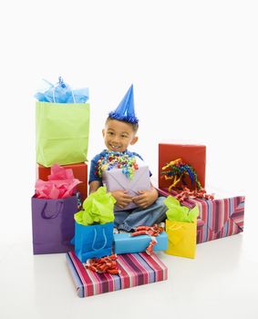 Asian boy wearing party hat sitting with pile of wrapped presents.