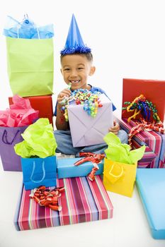 Asian boy wearing party hat sitting with pile of wrapped presents.