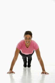 Mid adult multiethnic woman wearing pink exercise shirt doing pushups while looking at viewer and smiling.