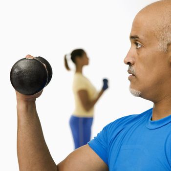 Close up of mid adult multiethnic man exercising using dumbbells with mid adult multiethnic woman in background.